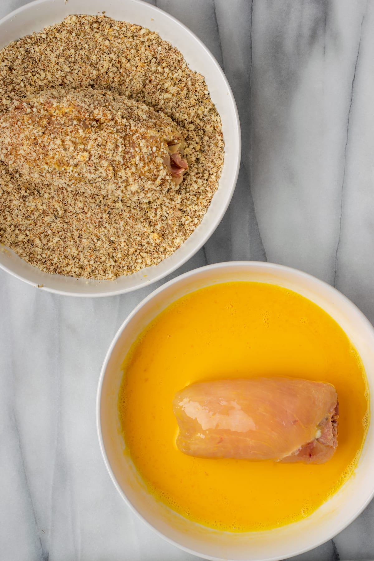 A bowl of breading and a bowl of beat eggs, both with a rolled up chicken breast in them