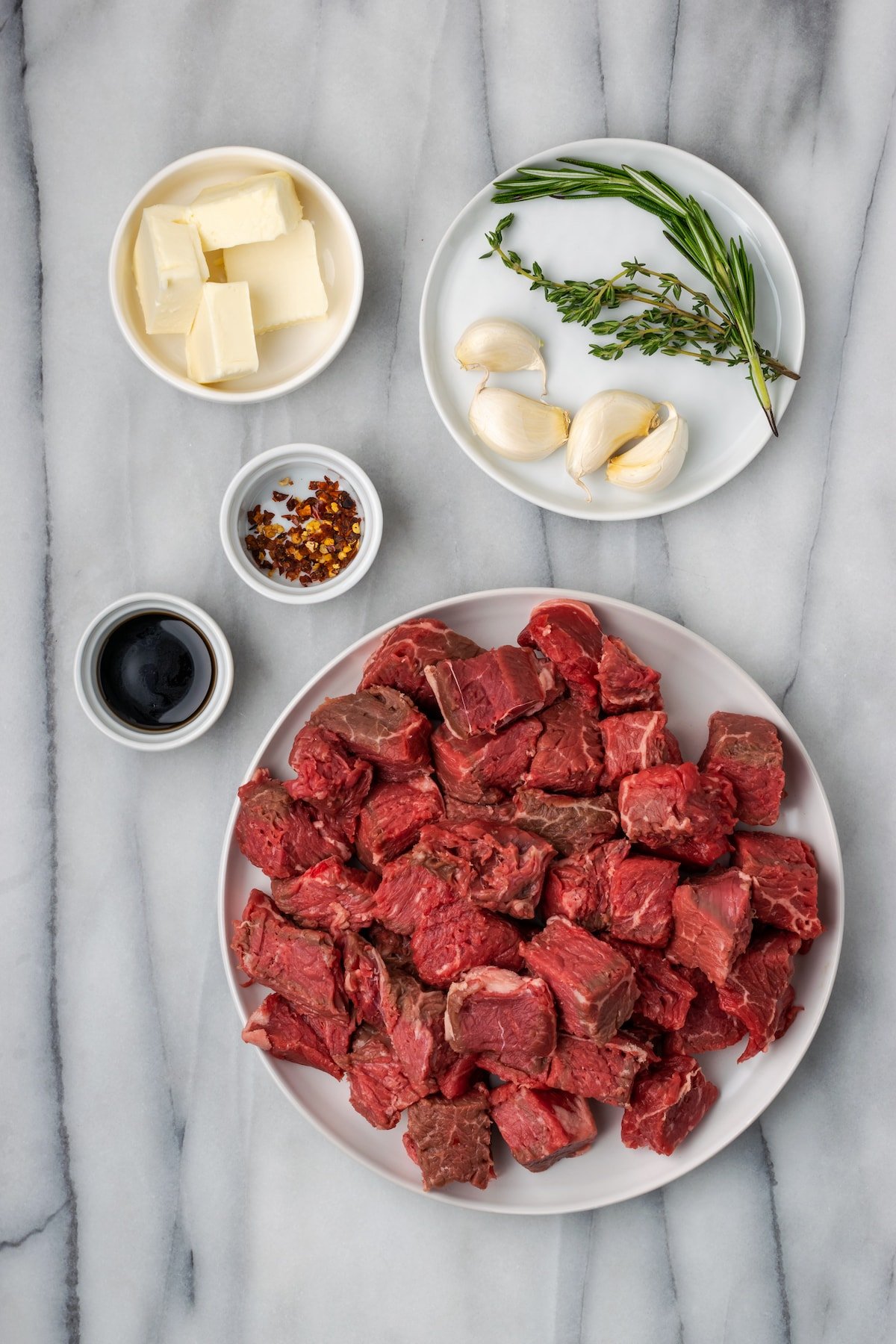 Overhead view of the ingredients needed for garlic butter steak bites: a plate of raw steak cubes, a bowl of butter, a bowl of soy sauce, a bowl of salt, pepper, and chili flakes, and a plate with garlic, fresh rosemary, and fresh thyme