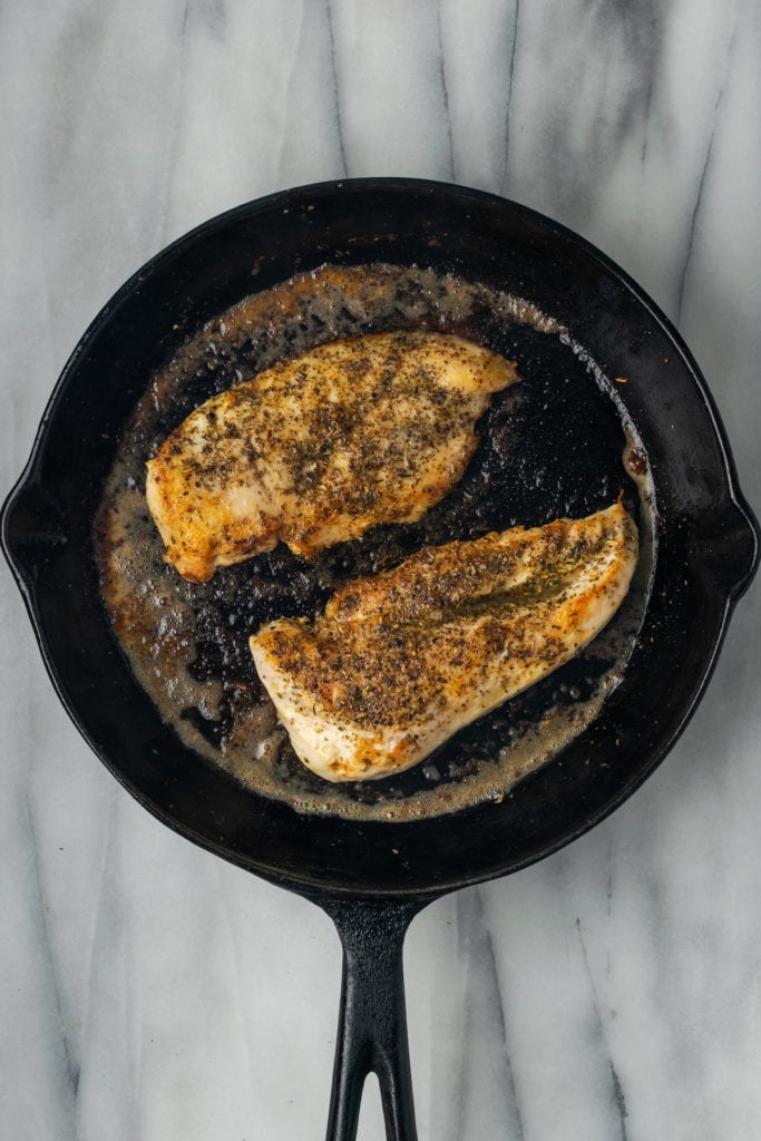 A skillet with two chicken breasts in it, with the tops seared