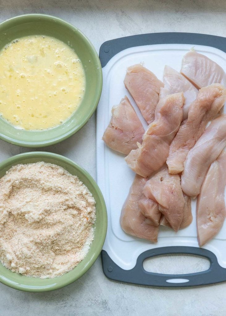 a cutting board with chicken tenders, and a breading station in green bowls