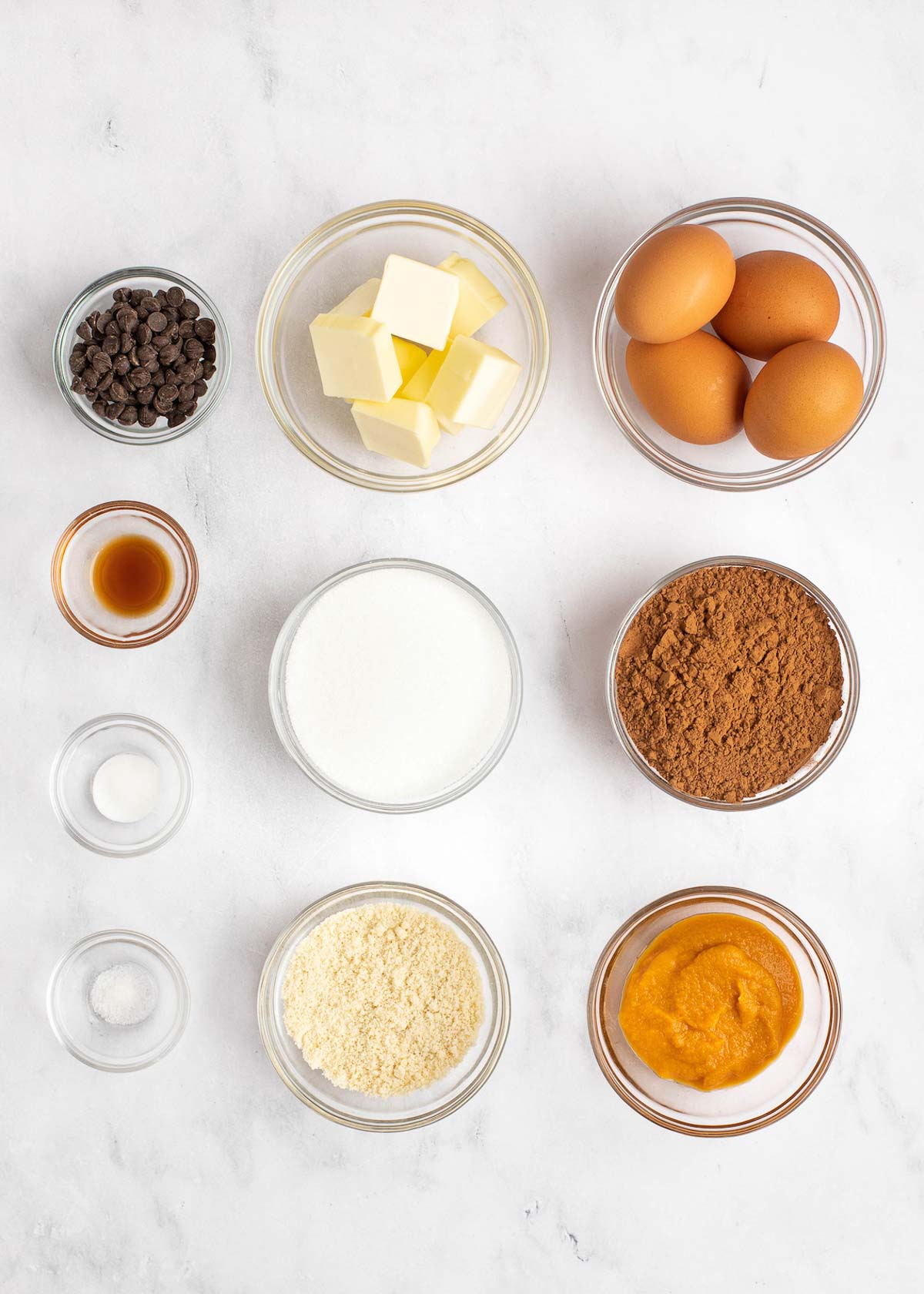 Overhead view of the ingredients needed for keto brownies: a bowl of eggs, a bowl of butter, a bowl of dark chocolate chips, a bowl of cocoa powder, a bowl of monkfruit sweetener, a bowl of pumpkin puree, a bowl of almond flour, a bowl of vanilla extract, a bowl of salt, and a bowl of baking soda