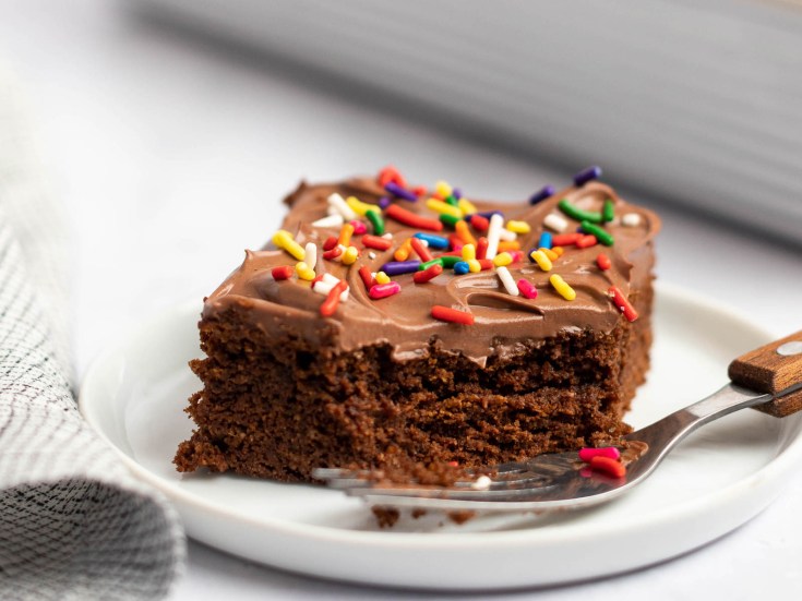 A piece of chocolate cake on a plate, covered with chocolate frosting and sprinkles, with a fork in front of it with a bite of cake on it