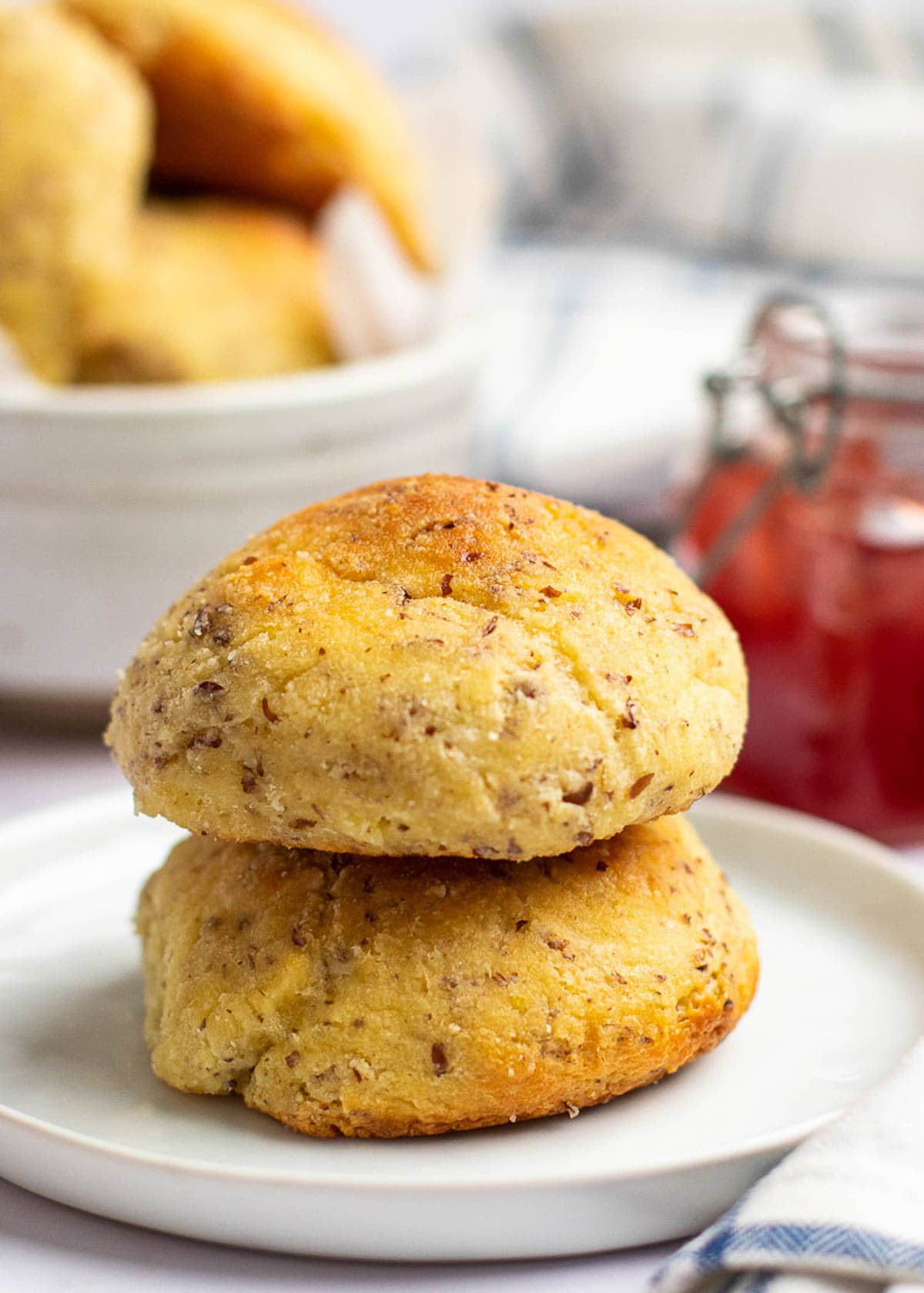 Two dinner rolls on top of each other on a plate, with a bowl of rolls and a jar of jam in the background