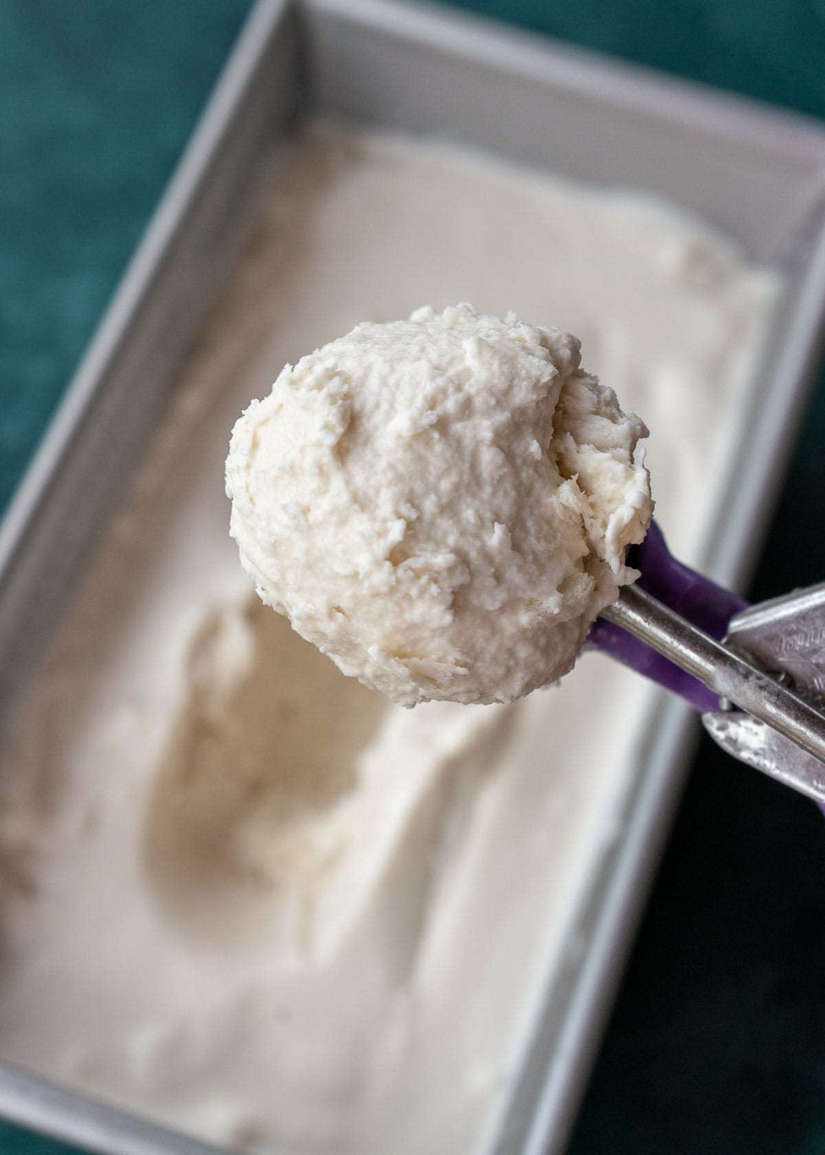 An ice cream scoop full of vanilla ice cream, with a loaf pan full of ice cream in the background