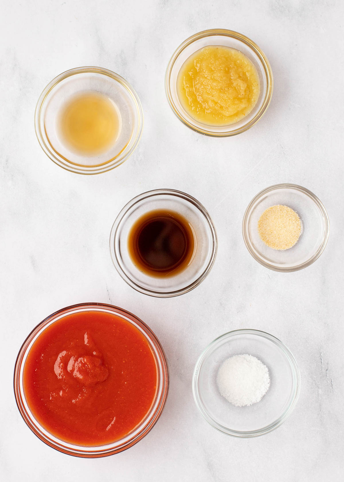 Overhead view of the ingredients needed for meatloaf sauce: a bowl of tomato sauce, a bowl of applesauce, a bowl of Worcestershire sauce, a bowl of apple cider vinegar, a bowl of salt, and a bowl of onion powder