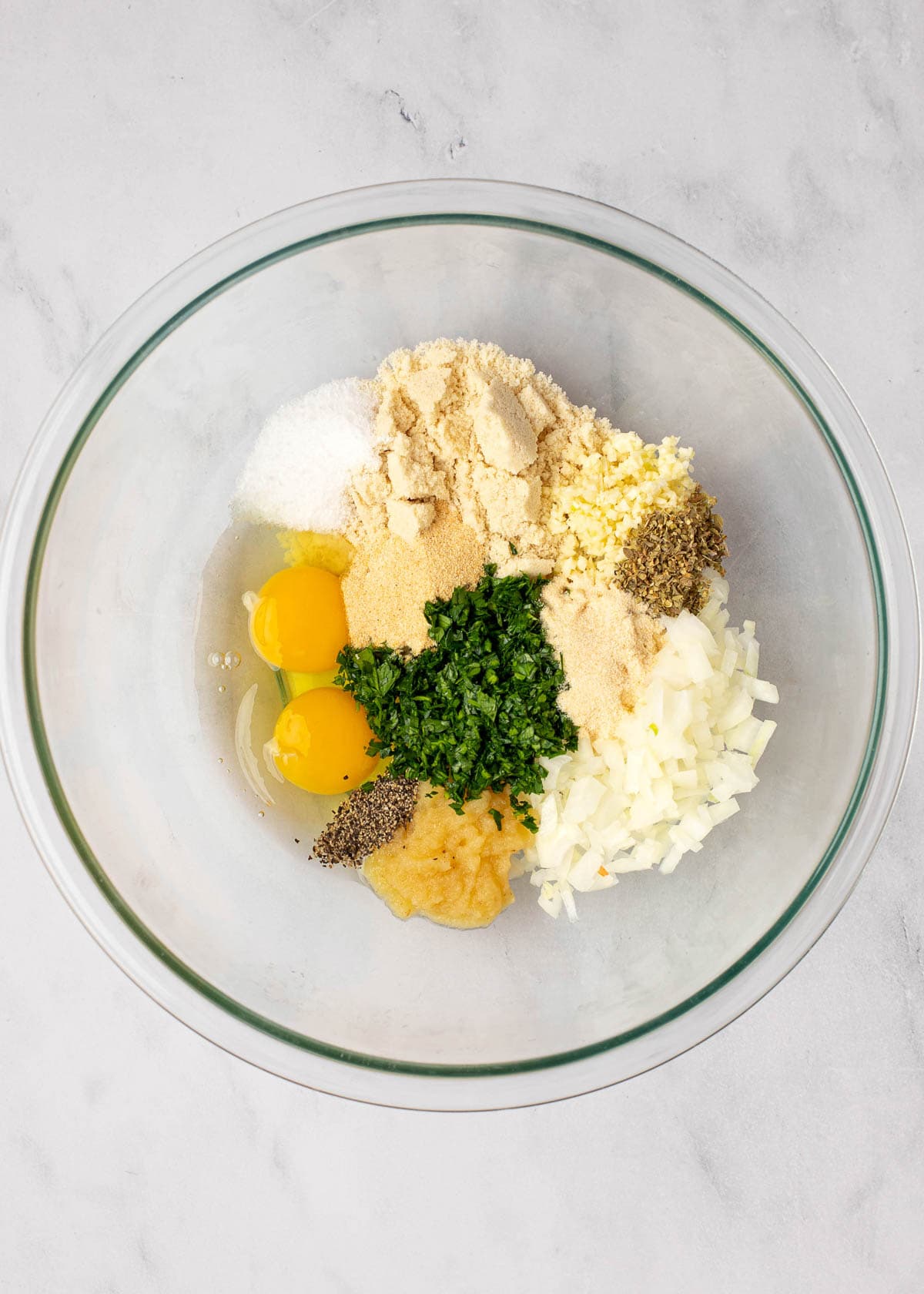 Overhead view of a mixing bowl with almond flour, onions, garlic, parsley, salt, pepper, arlic powder, onion powder, dried oregano, and eggs in it