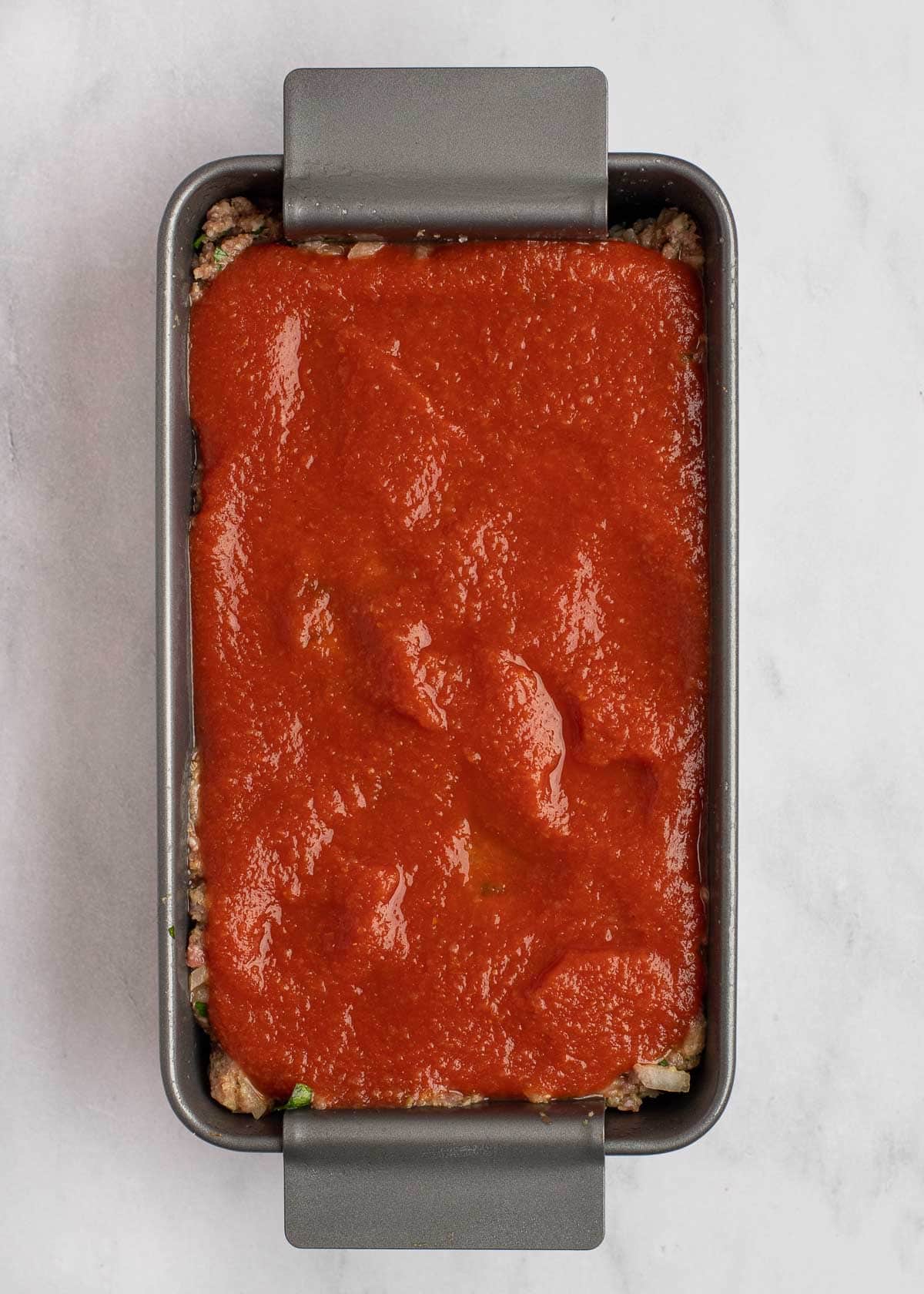 Overhead view of an unbaked meatloaf in a loaf pan covered in sauce