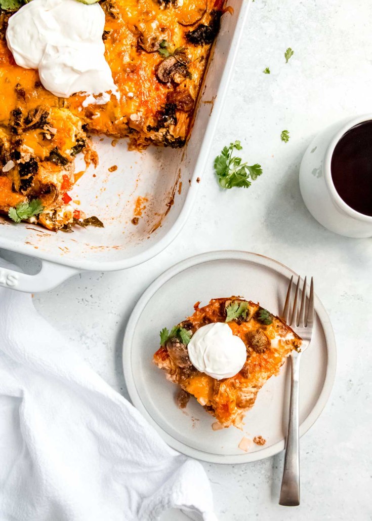 overhead shot of white table. Breakfast casserole baking dish is missing one piece; piece is garnished with sour cream and placed on a plate with a fork.